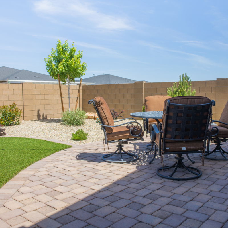 The Kann Residence boasts a well-maintained backyard featuring a paved patio area with a round table and four brown cushioned chairs. Enclosed by a tall brick wall, the yard includes a small tree, ornamental plants, and gravel landscaping. The clear blue sky completes the serene ambiance.