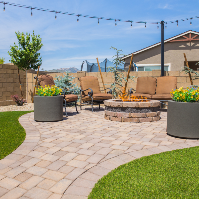 The Kann Residence&#039;s backyard patio features a fire pit surrounded by four cushioned chairs. Large potted plants with yellow and green flowers are placed nearby. String lights are hung overhead, and there&#039;s a trampoline and a house in the background. The ground is paved with bricks.