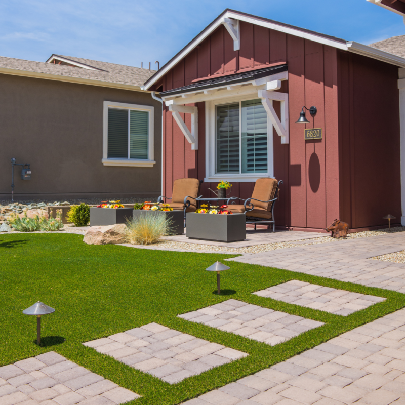 The Kann Residence is a charming house with maroon siding, white trim, and a neatly manicured front yard. The yard features a stone pathway with small lanterns leading to a patio with two cushioned chairs and a planter with flowers. Adjacent is a beige house with similar landscaping.