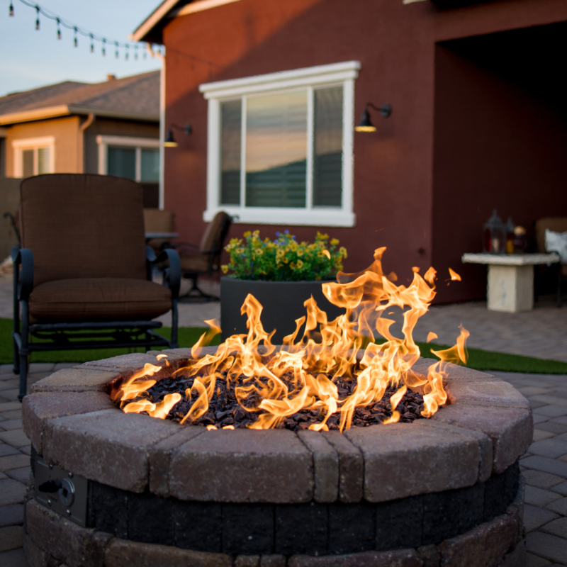 A warm, inviting fire pit on a brick patio in the Kann Residence&#039;s backyard. Chairs surround the fire pit, and string lights hang above. In the background, there is a house with large windows and a well-maintained garden with blooming flowers.