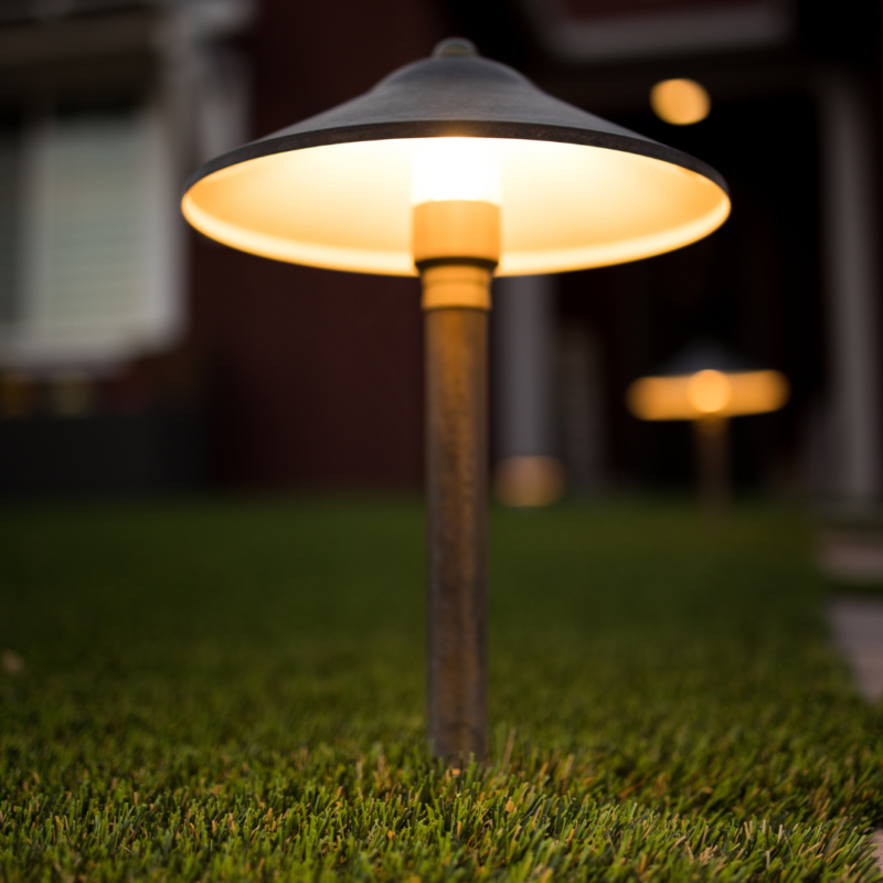 Close-up of a glowing pathway light with a black metal shade on neatly trimmed grass during dusk. Another identical light is blurred in the background, illuminating the path leading to the Kann Residence, with a partly visible window and door.