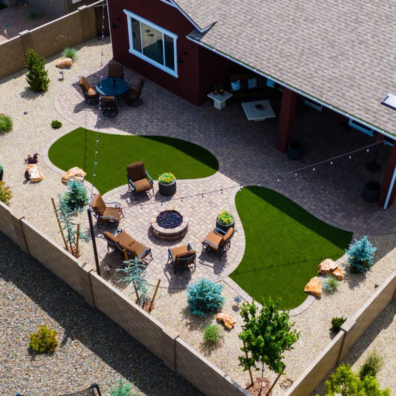 Aerial view of the Kann Residence&#039;s backyard featuring a patio with small patches of artificial grass. The patio boasts a round arrangement of chairs around a fire pit, with outdoor string lights, potted plants, and small trees enhancing the landscaped area.