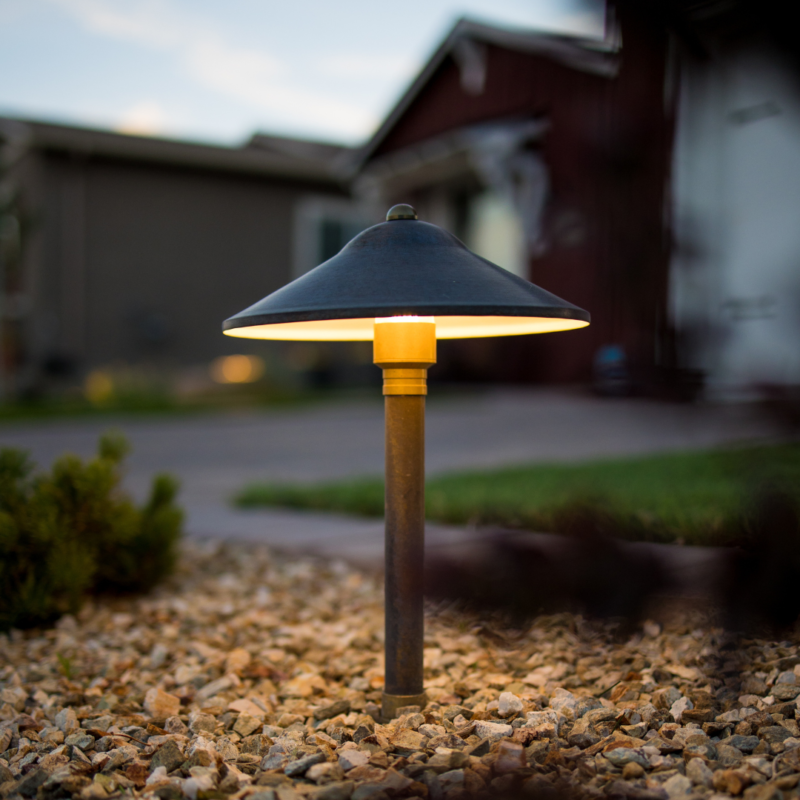 A small garden pathway light with a mushroom-shaped shade gently glows at dusk, illuminating the surrounding gravel at the Kann Residence. In the blurry background, there are houses and a driveway. The light casts a warm ambiance on the rocky ground.