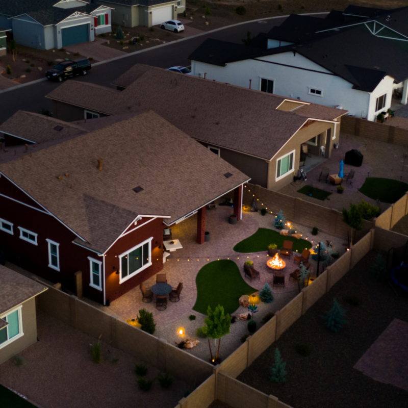 Aerial view of the Kann Residence&#039;s modern suburban backyard at dusk, featuring a cozy seating area with string lights and a fire pit. Surrounding houses and neatly landscaped lawns are visible, creating a serene neighborhood atmosphere.