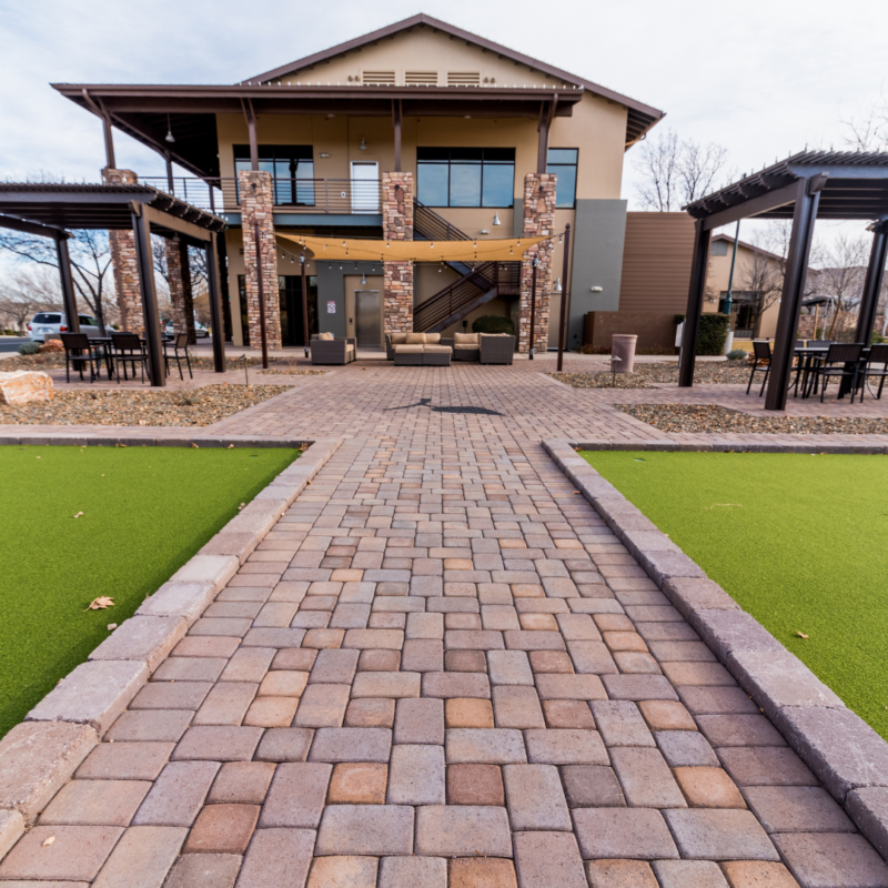 A modern two-story building, known as the StoneRidge Community Center, with stone pillars and balconies stands at the end of a paved walkway. The walkway is flanked by green artificial grass areas and shaded seating areas with tables and chairs. The sky above is clear.