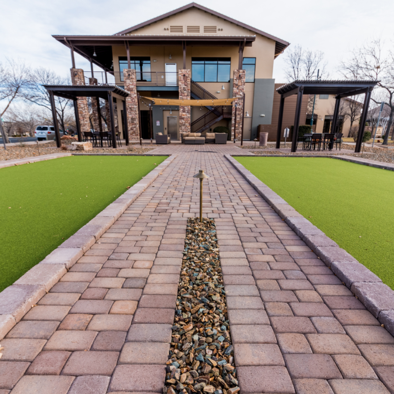 A modern two-story building with a stone facade and covered patios, the StoneRidge Community Center boasts a welcoming pathway with stone pavers and a landscaped strip of rocks leading to the entrance. Flanking the path are green synthetic grass areas, complemented by sparse trees dotted around.