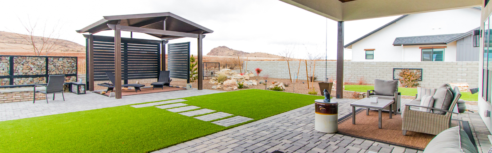 A modern backyard features a covered patio with seating, a paved area with steps leading to an open gazebo, a lawn with artificial grass, and a rock garden. In the background are hills, a short retaining wall, and a neighboring house.