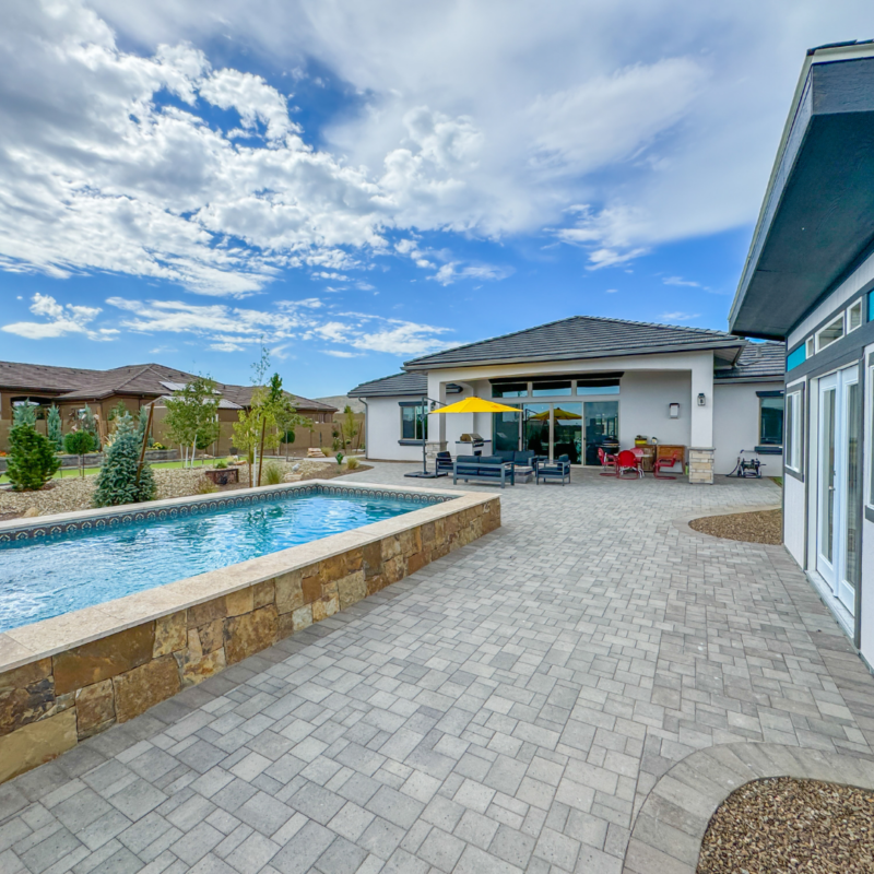 A modern backyard at the Hutchinson Residence features a stone-tiled patio with a rectangular swimming pool bordered by stone walls. Nearby, there&#039;s outdoor seating with a yellow umbrella. The house has large windows overlooking the pool and a bright, partly cloudy sky in the background.