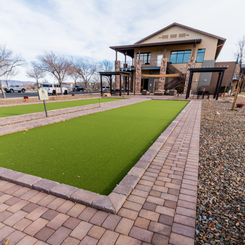 A modern house in the StoneRidge community boasts a large patio and second-floor balcony, featuring a bocce ball court in the backyard. A manicured lawn is flanked by paver walkways, and the area is surrounded by gravel landscaping with trees and parked vehicles in the background.