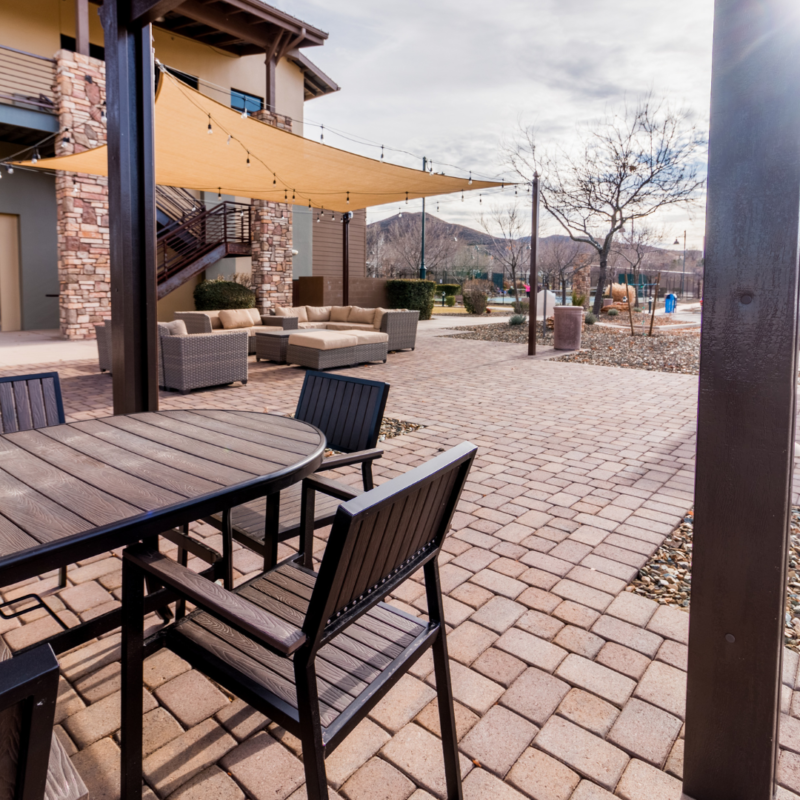 Outdoor patio area with brick flooring at the StoneRidge Community Center, featuring a dark wooden table and chairs in the foreground. Behind, beige cushioned sofas rest under a tan sunshade, flanked by trees and stone-accented buildings. Distant mountains and a cloudy sky complete the backdrop.