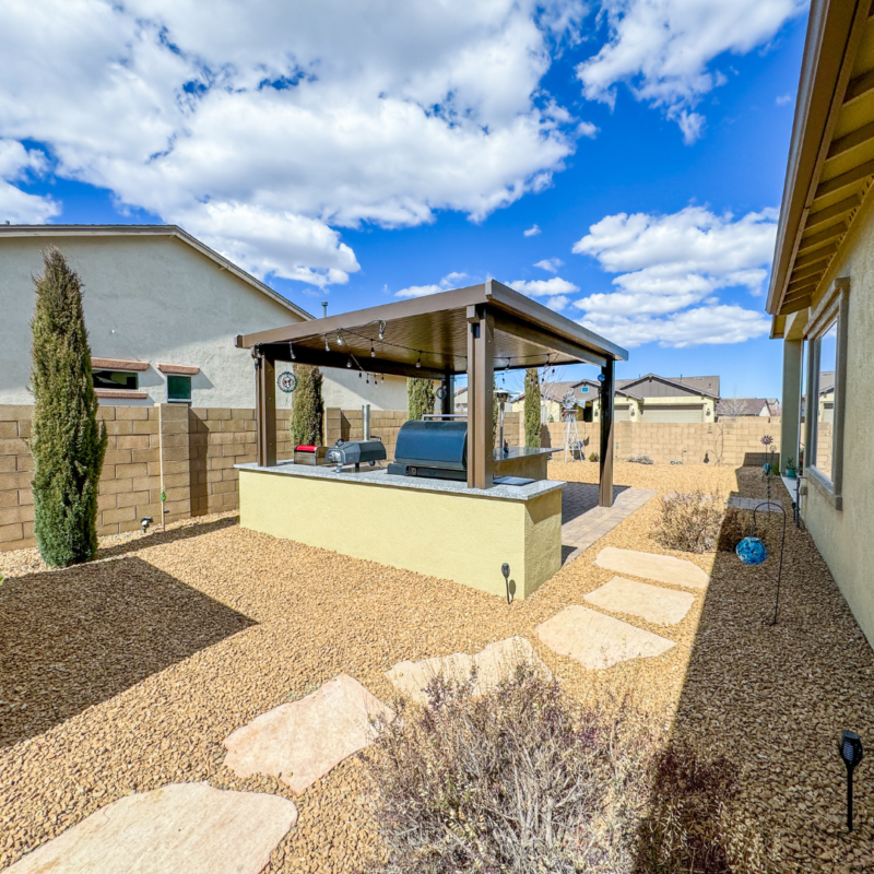 The Sutton Residence features a backyard with a covered pergola over an outdoor kitchen area complete with a grill. Gravel ground surrounds the pergola, accentuated by a few shrubs and stepping stones leading from the house. Above, the sky is blue with scattered clouds.