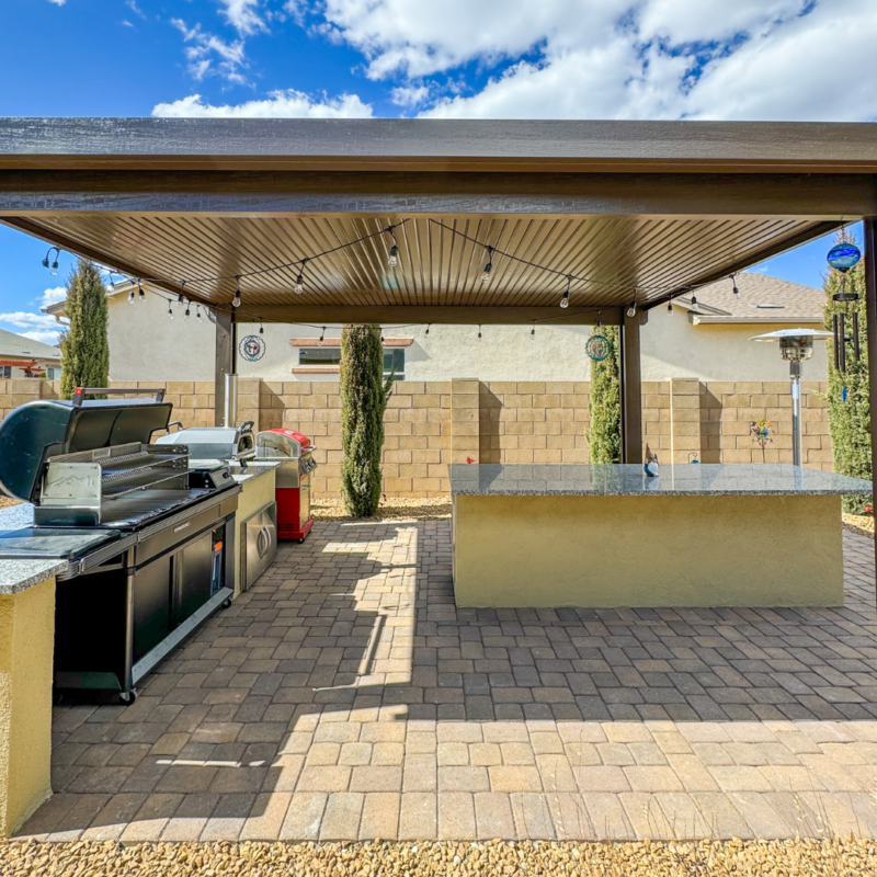 A spacious outdoor kitchen at the Sutton residence features a pergola covering a grill, sink, and countertop on a brick-paved patio. The area is surrounded by a low brick wall, with tall, thin trees lining it. The sky is clear with only a few clouds drifting by.