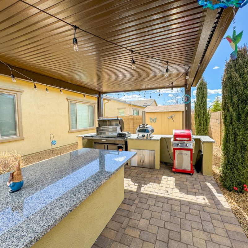 The Sutton Residence features a covered outdoor kitchen with a granite countertop, stainless steel appliances including a grill and silver pizza oven, and a red grill. Hanging string lights adorn the ceiling. The area is paved with bricks and bordered by beige walls and greenery.