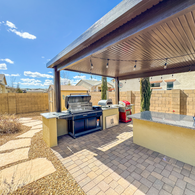 A backyard patio at the Sutton Residence with a covered outdoor kitchen featuring a grill, a red smoker, countertops, and storage cabinets. The area is paved with stone bricks and surrounded by a stone pathway, with a fenced wall and shrubs visible in the background.