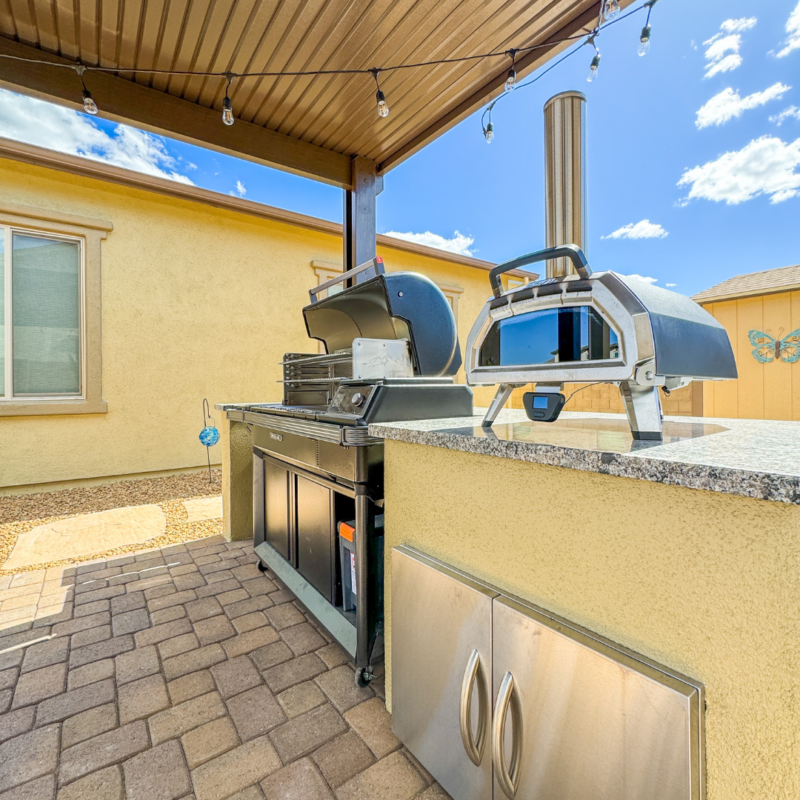 An outdoor kitchen at the Sutton Residence features a grill and a pizza oven under a pergola. The setup includes a countertop, storage cabinets, and hanging string lights. The kitchen is on a patio next to a beige house, with a sunny, clear sky in the background.