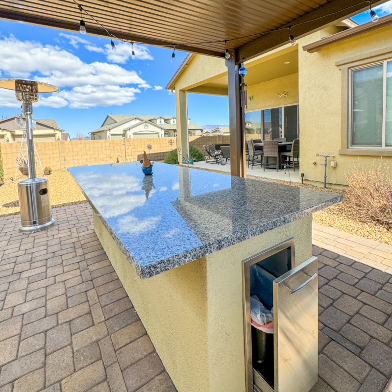 At the Sutton Residence, an outdoor kitchen with a granite countertop sits on a paved patio. The countertop features a cutout for a built-in trash bin. In the background, there are houses, a covered patio area with a dining table, and a propane heater. The sky is clear with a few clouds.