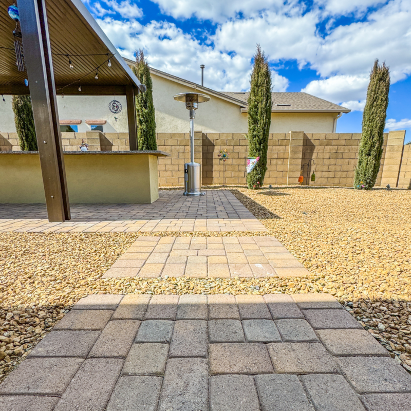 The Sutton Residence&#039;s backyard features a stone-paved pathway leading to a covered patio area with a roof, surrounded by gravel and tall, narrow trees planted along a stone wall in the background. A patio heater stands centrally, and the sky is partly cloudy.