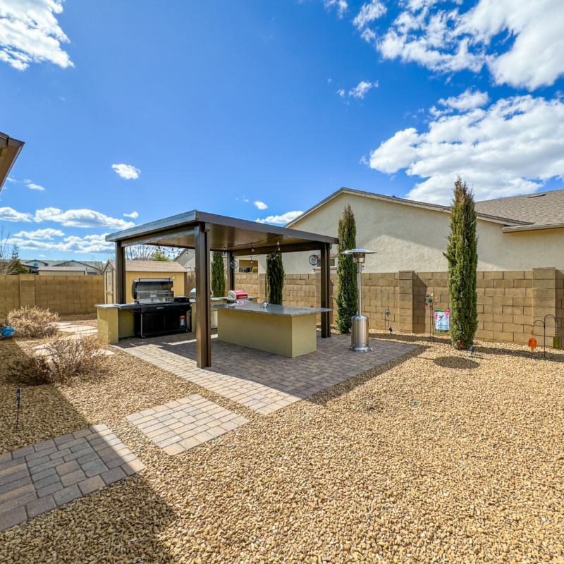 The Sutton Residence backyard features a covered outdoor kitchen with a built-in grill and countertop. The area is paved with stone tiles and surrounded by a gravel yard. Tall evergreen shrubs and a stone fence complete the space, all under a bright, partly cloudy sky.