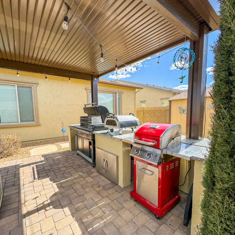 A covered outdoor kitchen at the Sutton Residence features a built-in grill, smoker, and a bright red barbecue grill on a stone countertop. This elegant setup is situated on a paved patio with string lights hanging from the roof, adjacent to two closed windows on the beige house wall.