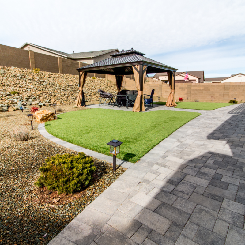 The Vaughn Residence features a backyard with a paved stone path leading to a gazebo housing a patio table and chairs on a grassy lawn. Decorative shrubs and a rock garden border the path. A stone wall encloses the area with houses visible in the background under a clear sky.