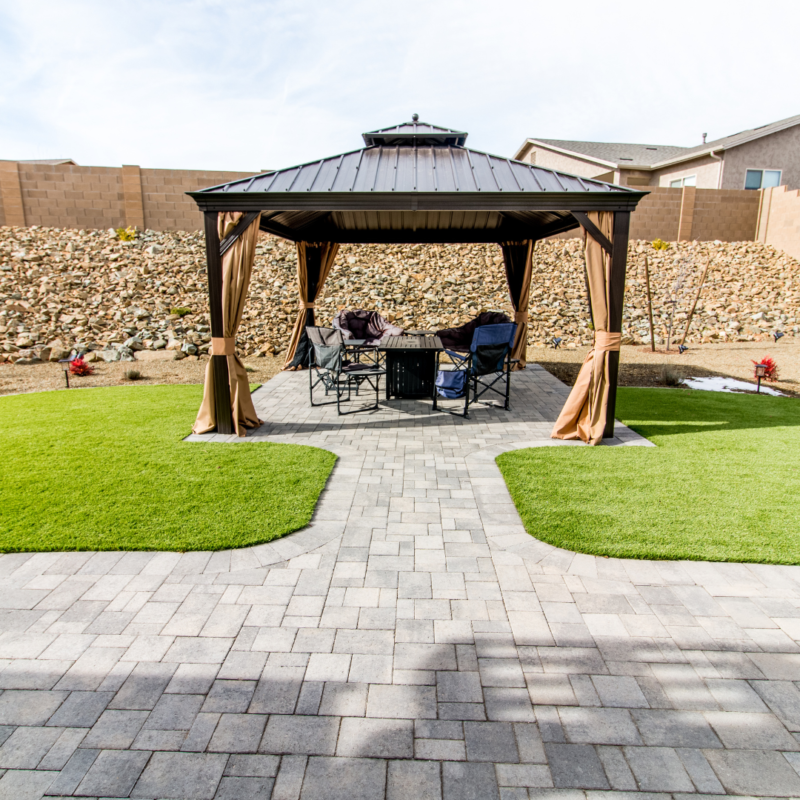 A backyard with a paved pathway leading to a gazebo. Under the gazebo at the Vaughn Residence, there is a set of outdoor furniture, including chairs and a table. The surrounding lawn and landscaping are well-maintained, with a stone wall and neighboring houses in the background.