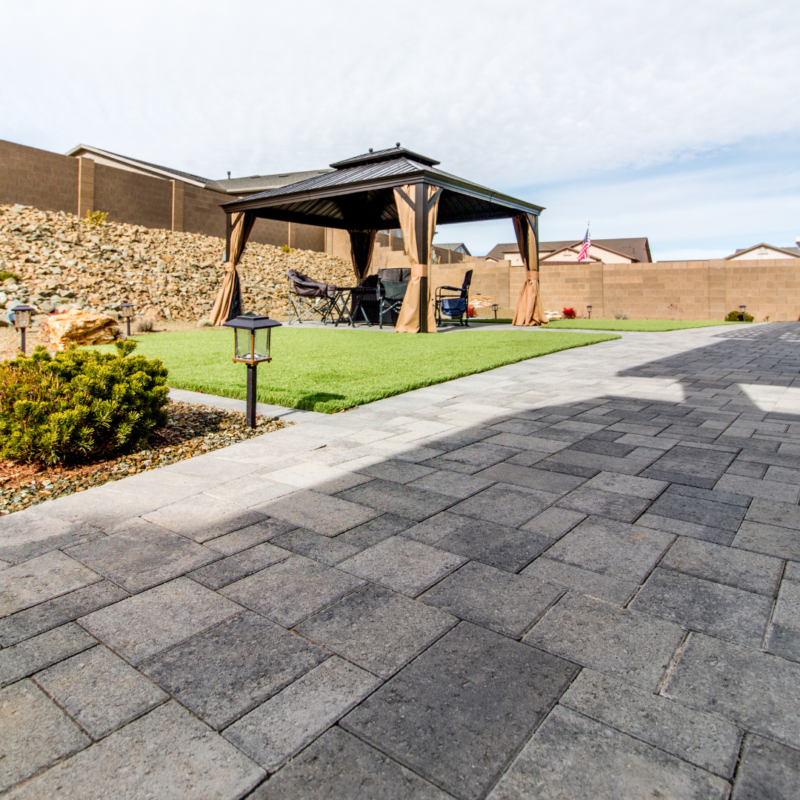 A backyard at the Vaughn Residence features a paved stone patio leading to a covered gazebo. Inside the gazebo are a table and chairs. The area is surrounded by a rock wall and a small patch of green grass. Landscape lights line the path, with an American flag visible in the background.