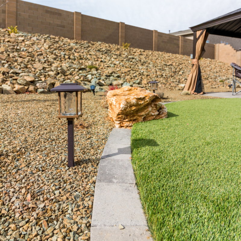 A backyard scene at the Vaughn Residence features a small garden with a mix of green artificial grass and gravel. A pathway lined with lanterns separates the two areas. A brown gazebo with a covered roof is situated on the grass near the right side of the image.