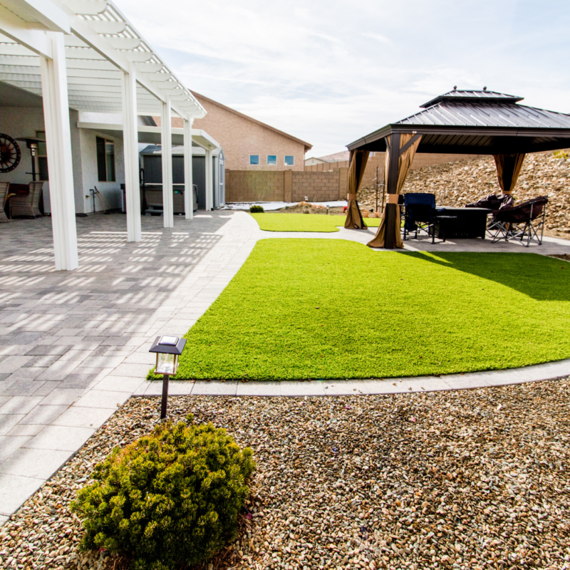 The Vaughn Residence boasts a backyard with a large patio featuring a white pergola, paving stones, and green artificial grass. To the right is a gazebo with outdoor furniture. The yard is bordered by a low stone wall and gravel. A small shrub and solar lamp adorn the foreground.