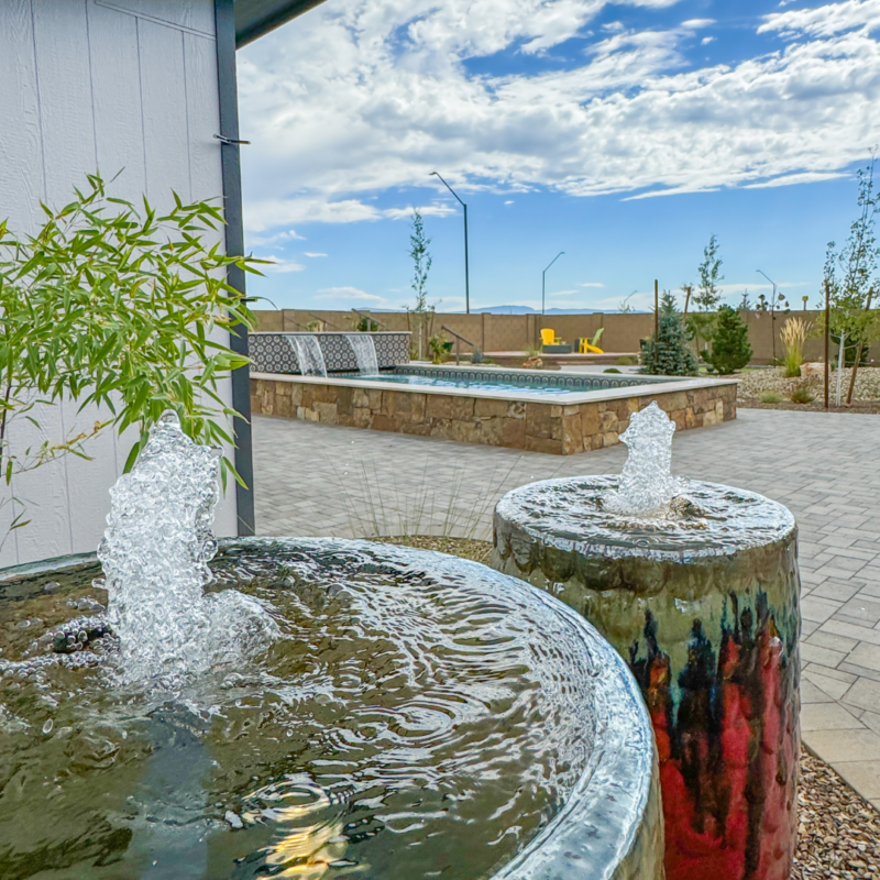 A serene outdoor setting at the Hutchinson Residence features two water fountains in large ceramic pots. Behind them is a brick patio area with a rectangular pool and stone wall. Some greenery is visible, and the sky is partly cloudy with mountains in the distance.