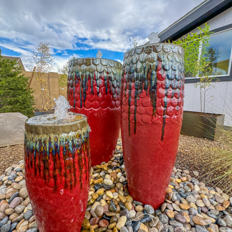 Three red and green ceramic water fountains stand among multi-colored pebbles in the garden of the Hutchinson Residence. Each fountain features cascading water flowing down its textured surface. In the background, there is a modern building and a clear sky with scattered clouds.