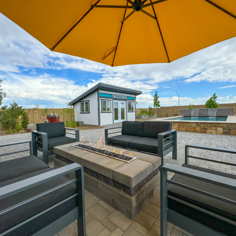 The Hutchinson Residence features an outdoor patio area with black cushioned chairs and a central fire pit table under a large yellow umbrella. A modern shed in the background is surrounded by a paved area and greenery, all set against a partly cloudy sky.