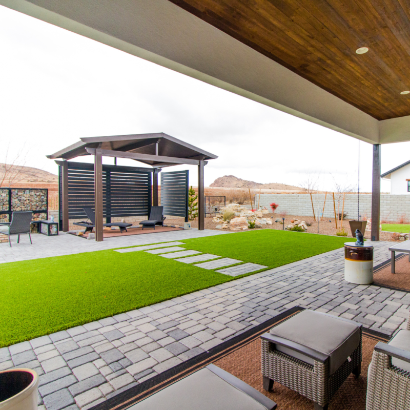 A modern backyard at the Lange residence features a covered patio with wicker furniture, leading to a green lawn with stepping stones. Beyond the lawn, there&#039;s a pergola with lounge chairs on a paved stone area. The scene is framed by a desert landscape with mountains in the background.