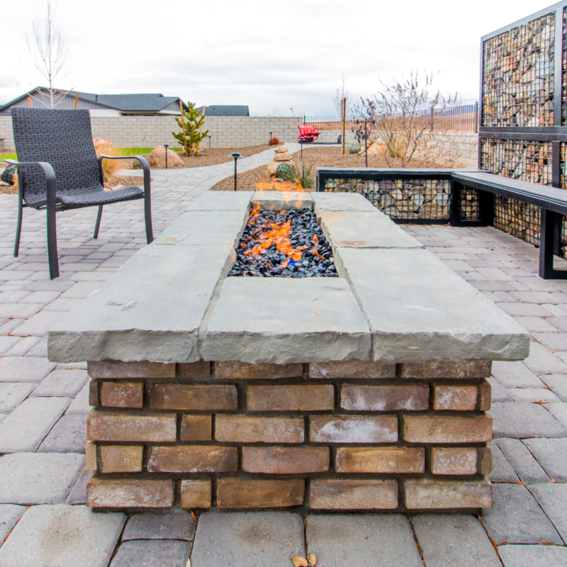 The Lange Residence boasts a modern outdoor patio featuring a rectangular fire pit with a stone and brick base. The fire pit is lit with a vibrant flame, surrounded by patio chairs, stone flooring, and a decorative rock-filled fence. In the background, trees stand tall against a cloudy sky.