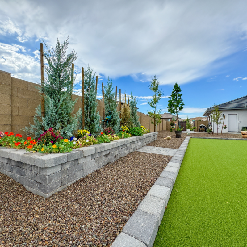 The Hutchinson Residence boasts a landscaped backyard with a neatly paved stone walkway, a raised flower bed brimming with colorful flowers and shrubs against a stone wall, and a lush green artificial turf area. In the background, the house stands under a cloudy blue sky.