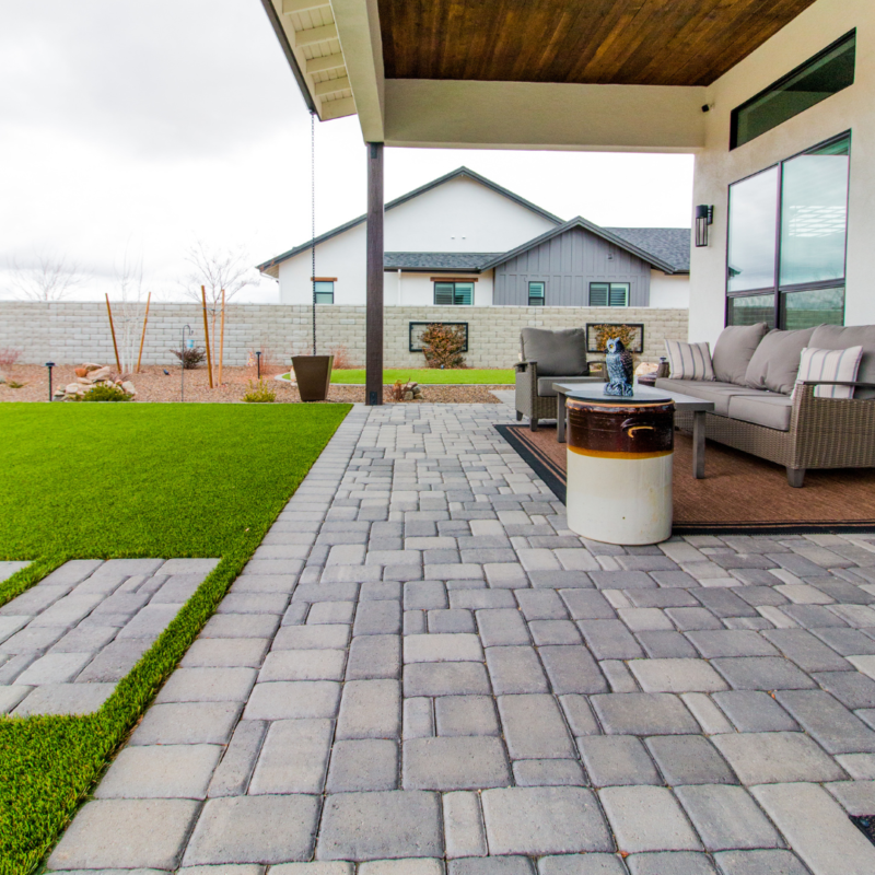 A covered patio area with cushioned seating and a small table overlooks the Lange Residence&#039;s backyard, featuring green artificial grass and a pathway made of gray pavers. Beyond the yard, the modern house stands against a cloudy sky, with sparse landscaping lining the fence.