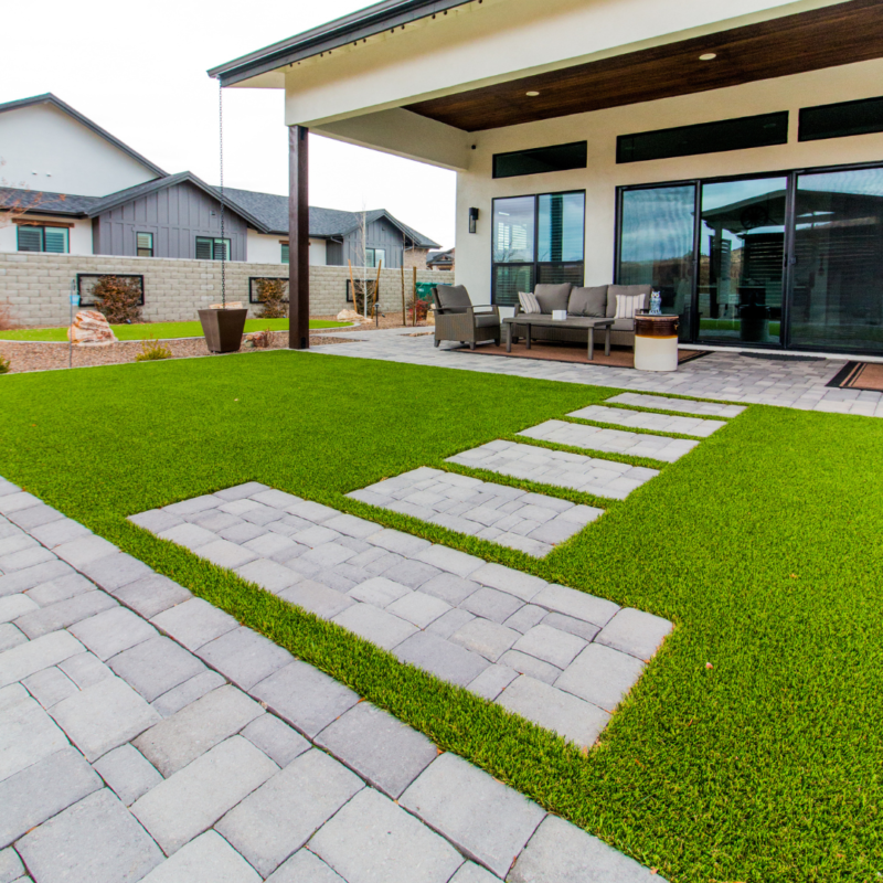 The Lange Residence boasts a modern backyard patio with a covered seating area furnished with a sofa set and table. The yard features a green lawn with a stone path and tiles, bordered by neighboring houses, along with a short brick wall and wooden fencing in the background.