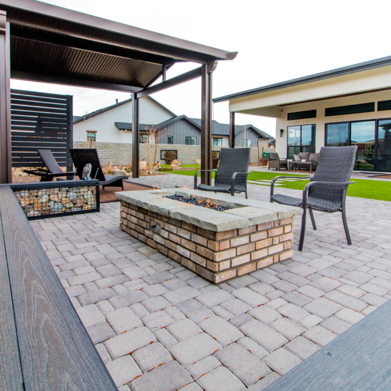 Outdoor patio area with cobblestone flooring features a rectangular brick fire pit in the center. At Lange Residence, two wicker lounge chairs surround the fire pit. A covered seating area with two chairs is in the background, adjacent to a modern building.