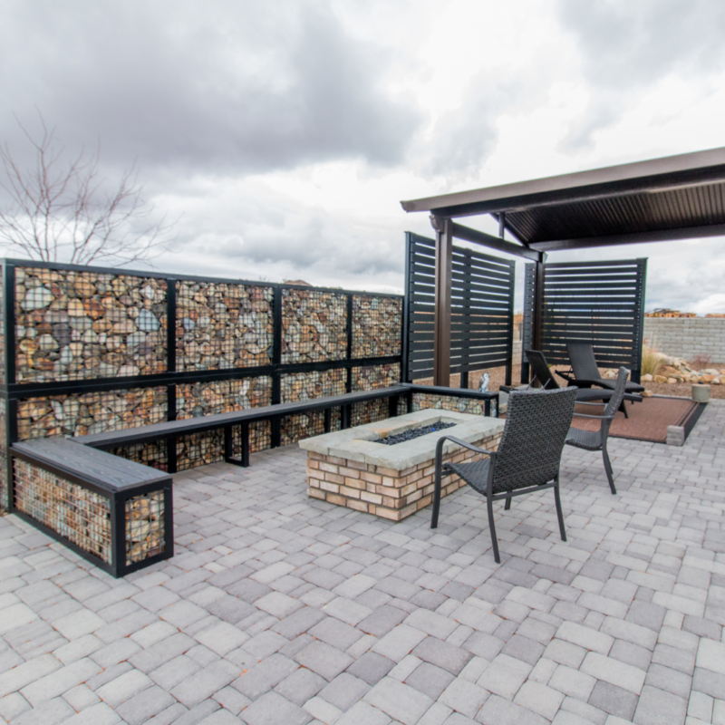 The Lange Residence boasts a modern patio featuring a brick fire pit, black metal chairs, and a sleek metal privacy screen. Decorative wire gabions filled with rocks form an eye-catching wall, while the gray brick paving extends to an overlook of the cloudy sky.