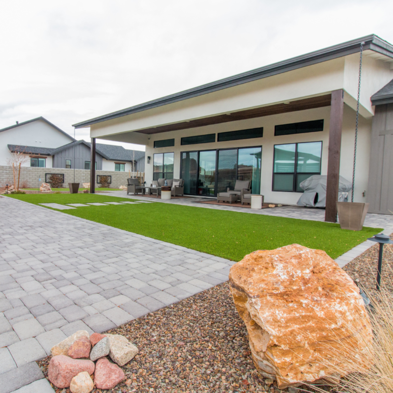 The Lange Residence is a modern house with a large covered patio featuring glass doors and windows. The backyard includes a green lawn, a paved area, and decorative rocks and plants. Nearby houses and a gray sky are visible in the background.