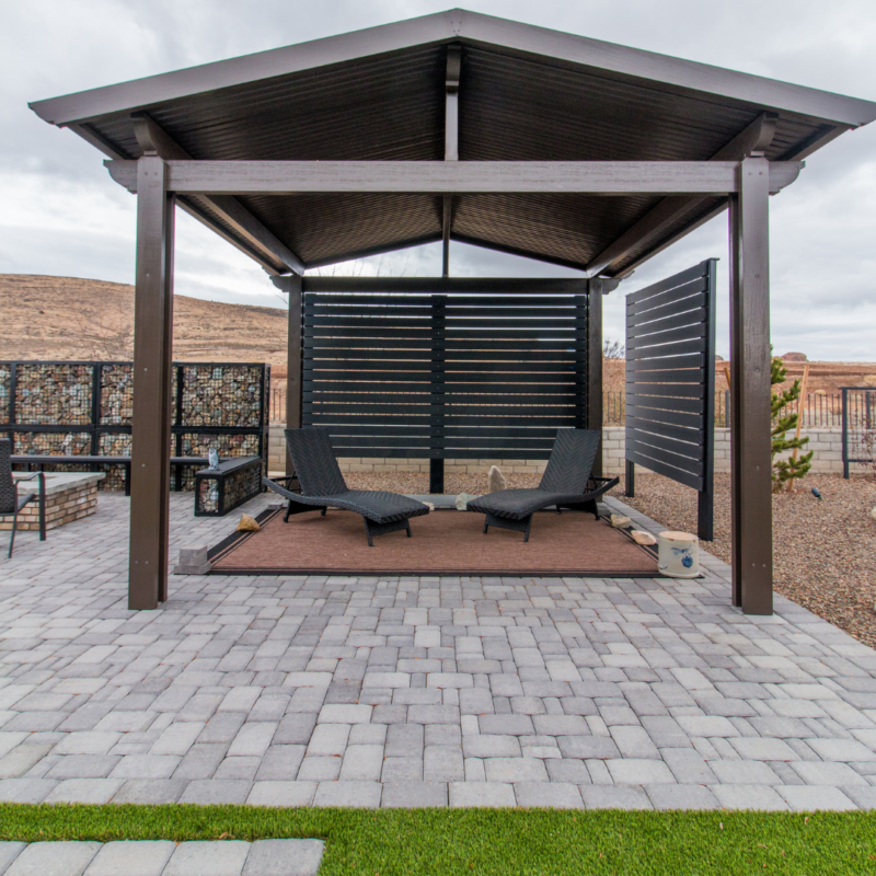 The Lange Residence features a modern outdoor gazebo with a metal roof, wooden slat walls on two sides, and two black reclining chairs on a small deck. The area is surrounded by a paved patio with green artificial grass in the foreground, set against a backdrop of a hilly, barren landscape.