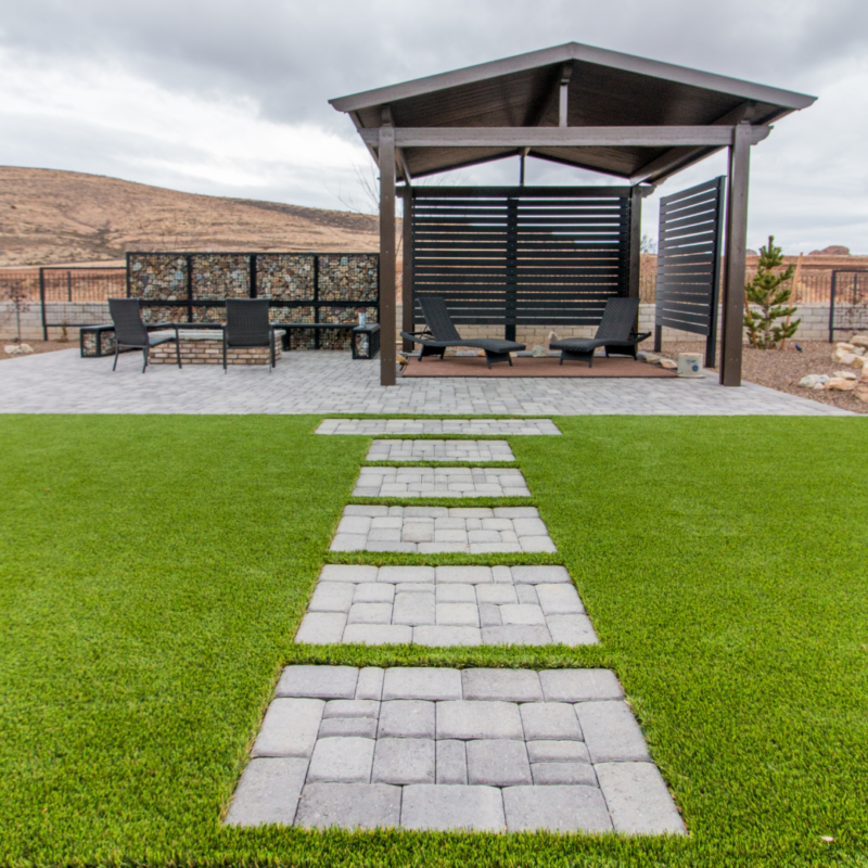 A well-maintained backyard at the Lange Residence features a paved stone pathway leading to a shaded gazebo. The gazebo has lounge chairs and a stone wall adjacent to it. The yard boasts neatly trimmed green grass and a scenic backdrop with hills and a cloudy sky.