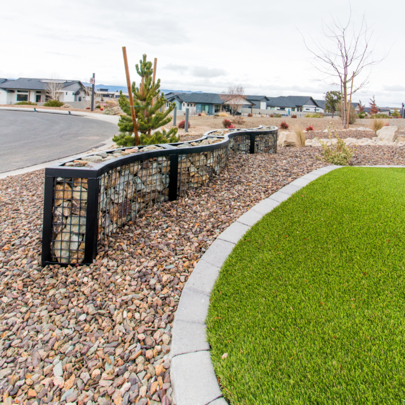 A landscaped yard at the Lange Residence features a curved, stone-filled gabion wall and lush green grass edged with gray stone edging. Surrounding the yard are houses, a paved street, and a leafless tree, with mountains faintly visible in the background under a cloudy sky.