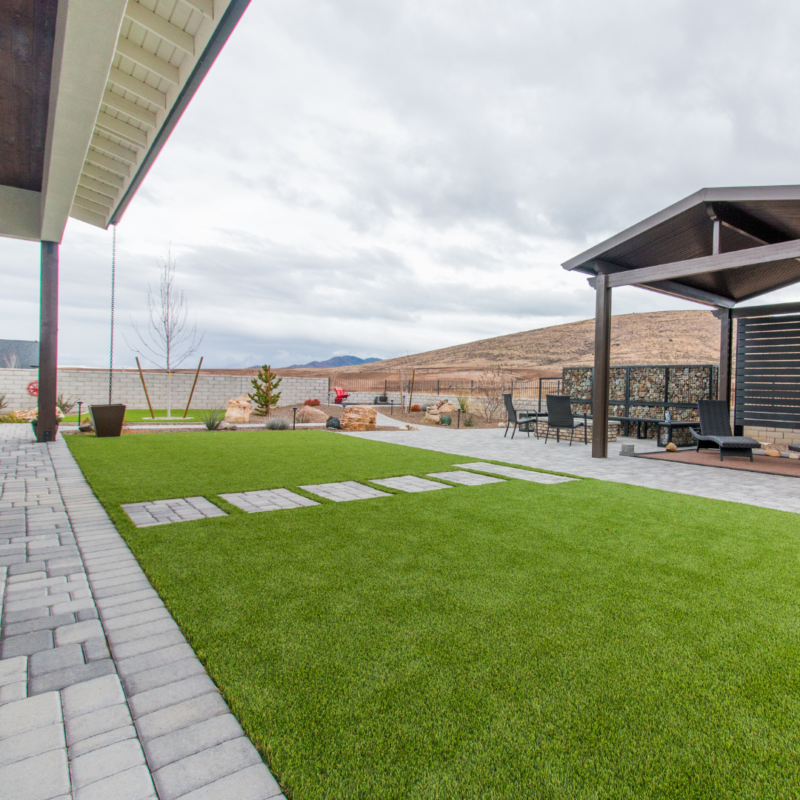 A modern residence with a neatly manicured lawn, stepping stones, a large patio area with outdoor furniture, and a pergola. The patio has pavers and is adjacent to a brick wall. The landscape, designed by Lange, features decorative plants and rocky areas with a hilly backdrop.