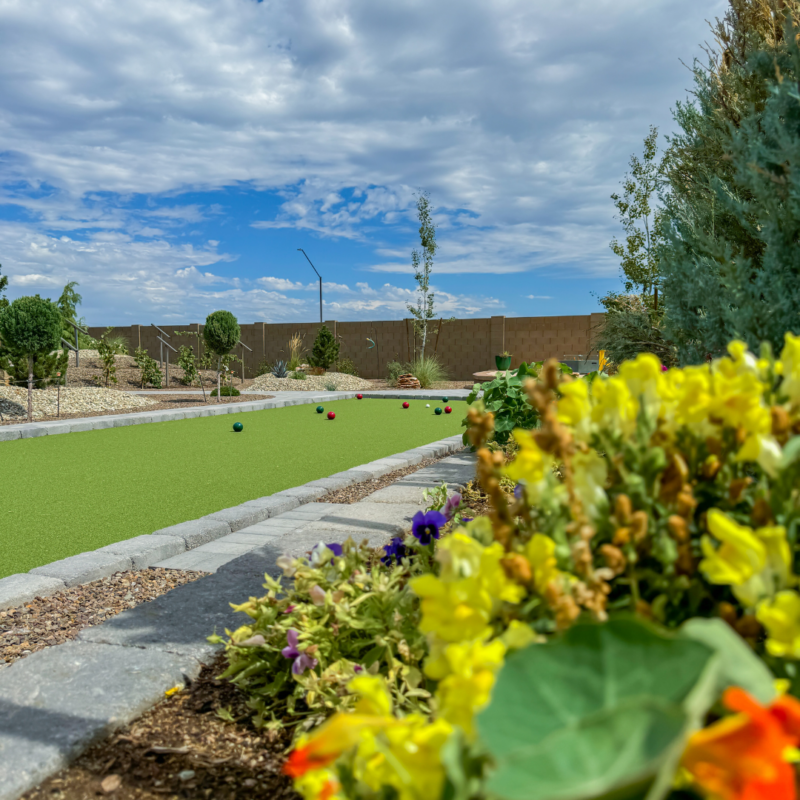 A bocce ball court under a partly cloudy sky graces the Hutchinson residence, with green grass surrounded by meticulous landscaping. Bright yellow and purple flowers adorn the foreground, while several bocce balls are scattered on the grass. Tall trees and a brown fence frame the serene scene in the background.