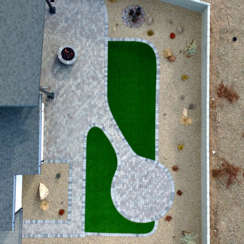 Aerial view of the Brown Residence backyard featuring a curved, green artificial lawn surrounded by gray brick paving. The yard includes a circular seating area with a fire pit, desert rock landscaping, and a modern house on the left side.