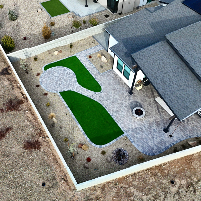 An aerial view of the Brown Residence backyard showcases a modern house with a tiled patio. The yard features artificial grass patches and decorative stone paths, a fire pit, and a few small plants and shrubs. A white fence borders the beautifully landscaped space.