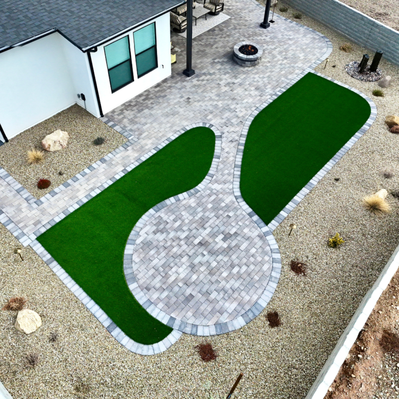 Aerial view of the Brown Residence&#039;s landscaped backyard with a paved patio extending from the house. The patio features light gray pavers and is surrounded by two kidney-shaped artificial grass areas. The edges are lined with gravel and a few desert plants and rocks.