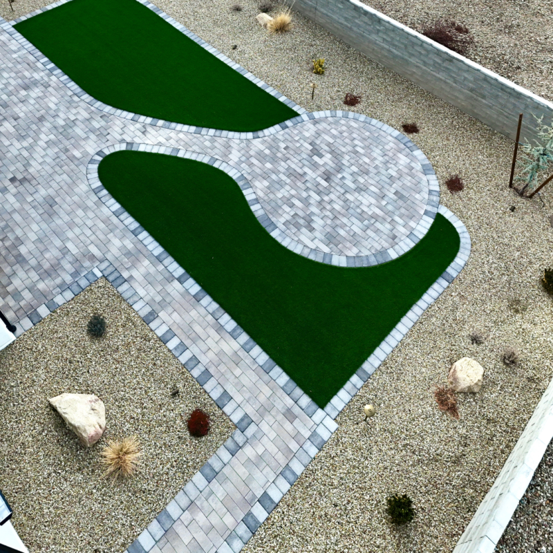 An aerial view of the Brown Residence&#039;s landscaped backyard features a paved pathway and patio area with curved edges, bordered by green artificial grass. The surrounding area is covered with small gravel, and there are a few scattered rocks and dry plants.