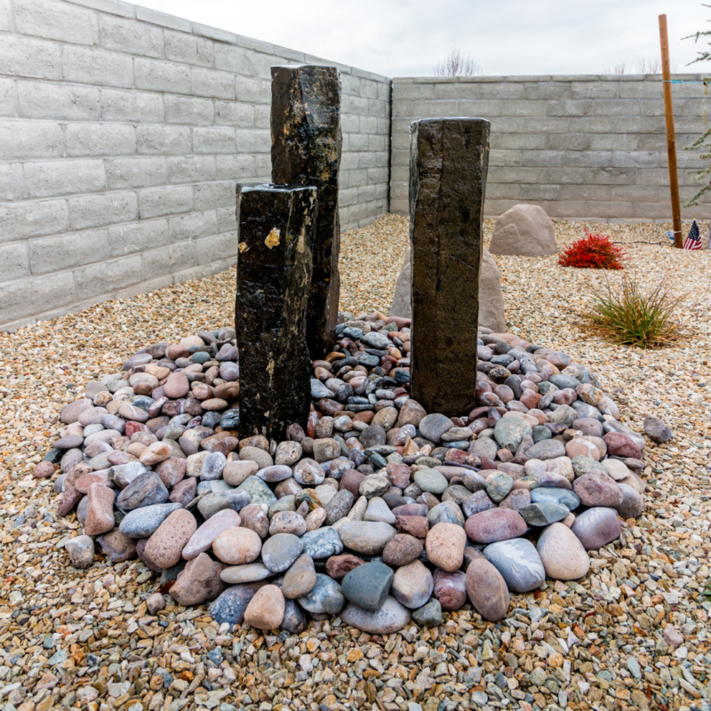 The Brown Residence boasts a minimalist garden featuring three vertical stone pillars within a circular arrangement of multicolored rocks. The ground is covered with gravel, and a stone wall encloses the space. Sparse vegetation accents the tranquil, zen-like setting.