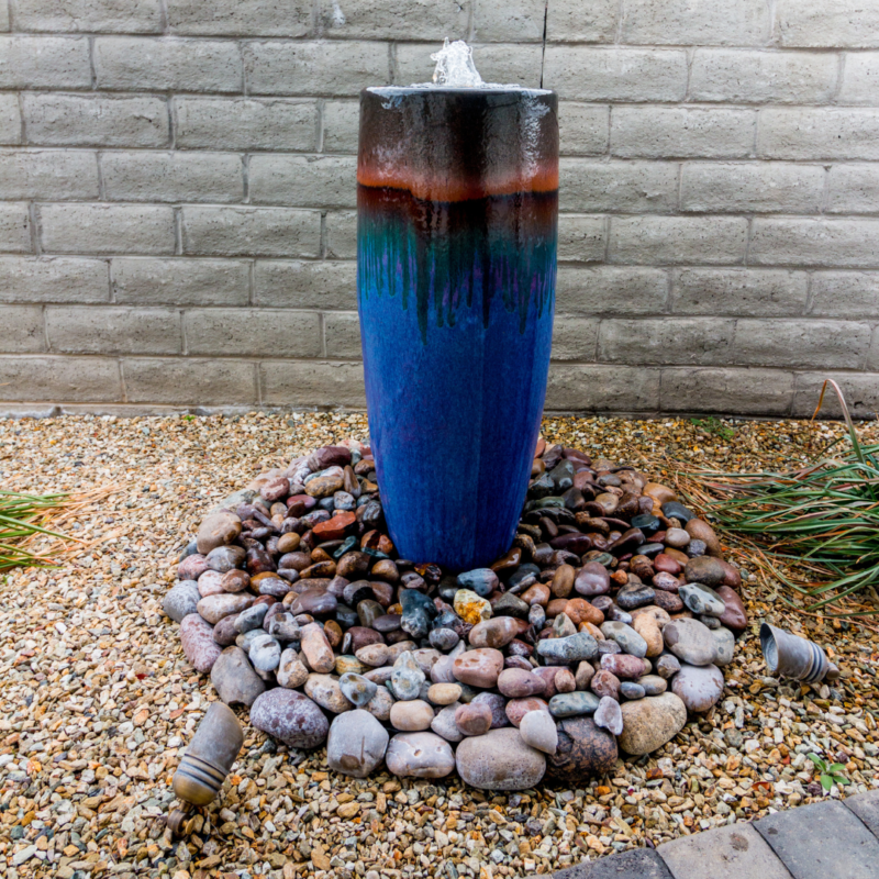 A tall, cylindrical blue ceramic water fountain serves as the focal point in a residence garden. Surrounded by a bed of colorful small stones on a gravel surface, water cascades gently from the top. Two garden lights are positioned on the ground, enhancing the serene ambiance of this brown-hued haven.